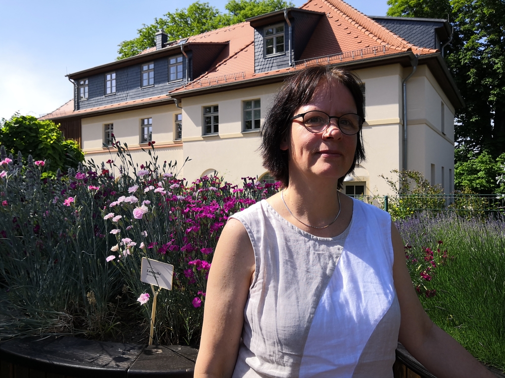 Pastorin Fourestier im Duftgarten, links bunte Blumen, im Hintergrund das Haus "Storchennest"