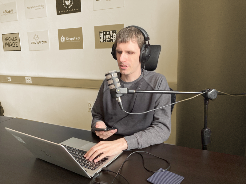 Dennis Westphal am Tisch vor einem Mikrofon mit Kopfhörer sitzend. In der Hand ein Phone, vor sich ein Laptop. An der Wand dahinter Logos der Geschäftsbereiche der Gesellschaft zur Entwicklung von Dingen, z.B. den Bereich Broken Image