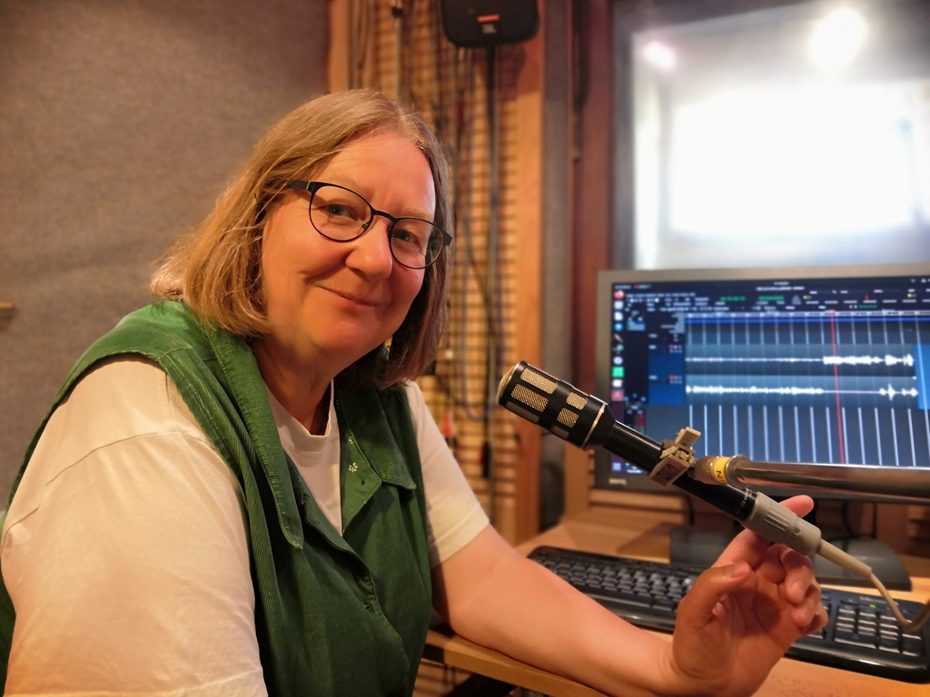 Dorothee Janssen schaut in die Kamera. Mit dem Finger zeigt sie zum Mikrofon. Im Hintergrund auf dem Bildschirm die OpenSource Audiobarbeitungssoftware Ardour