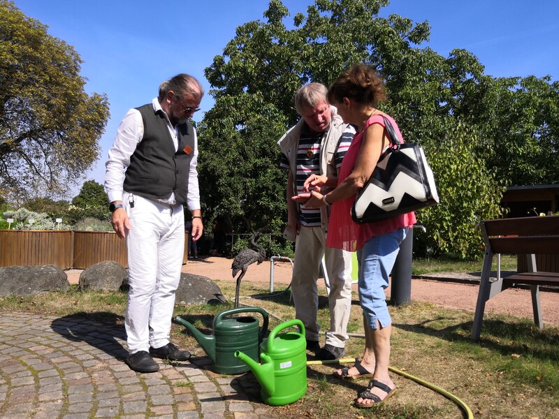 Einem taubblinden Besucher wird in die Hand gelormt während er vor zwei Gießkannen steht. Verwaltungsleiter Gerold Augart steht rechts und betreut die Aktion "Das große Baumbaden"