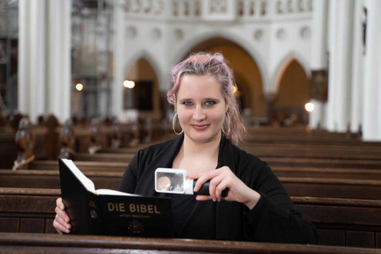 Eine junge Frau sitzt mit einer Leselupe und der Bibel in einer Bank einer großen Kirche, Bild DBSVT Friese