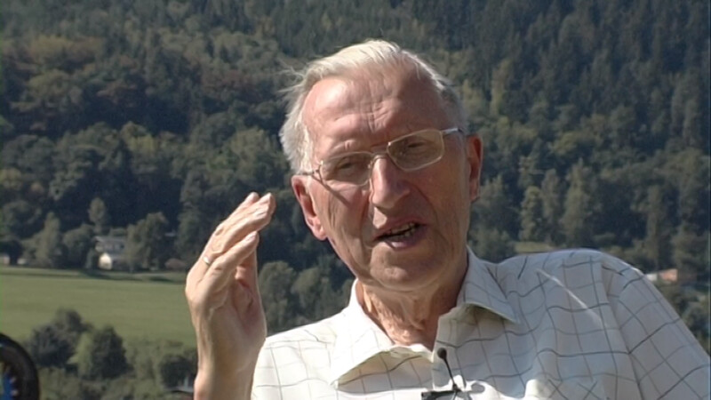 Pastor Uwe Holmer ist nah zu sehen. Eine Hand hebt er im Gespräch. Im Hintergrund ein Wald. Das Foto ist aus einem vieo, welches im Freien aufgenommen wurde.