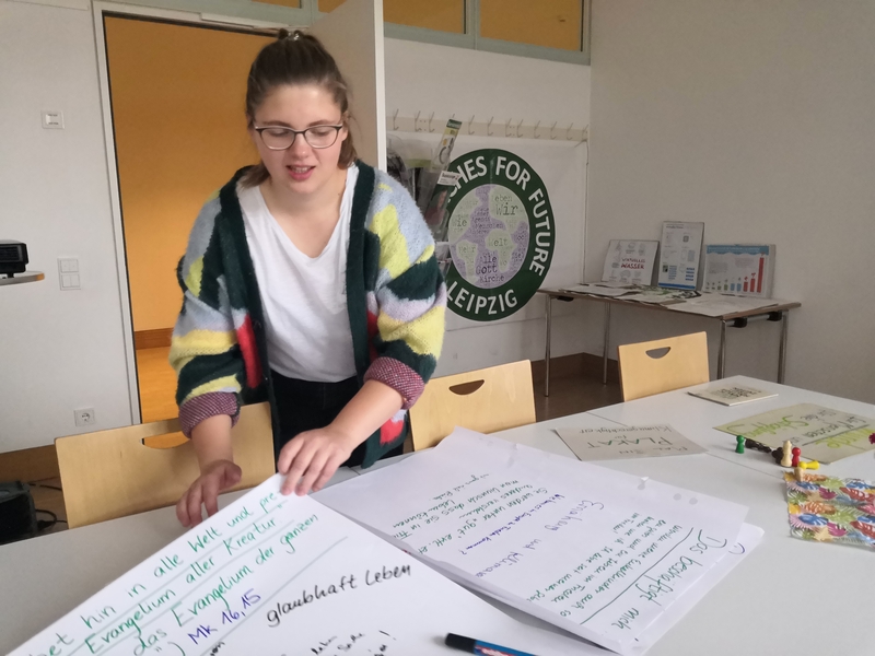 Michelle Schwarz, räumt Arbeitsmaterial eines Workshops auf. In der Hand hält sie ein großes Blatt auf dem die Schrift "Geht hin in alle Welt...glaubhaft leben" erkennbar ist. Im Hintergrund hängt das runde Logo "Churches for future"