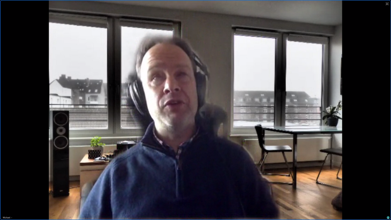 Foto: Das Gesicht von Michael Kuhlmann ganz nah mit Kopfhörer mit einem virtuellen Videokonferenzhintergrund (ein Wohnzimmer mit hellen Fenstern, Tisch, Stühle, große Lautsprecherbox)